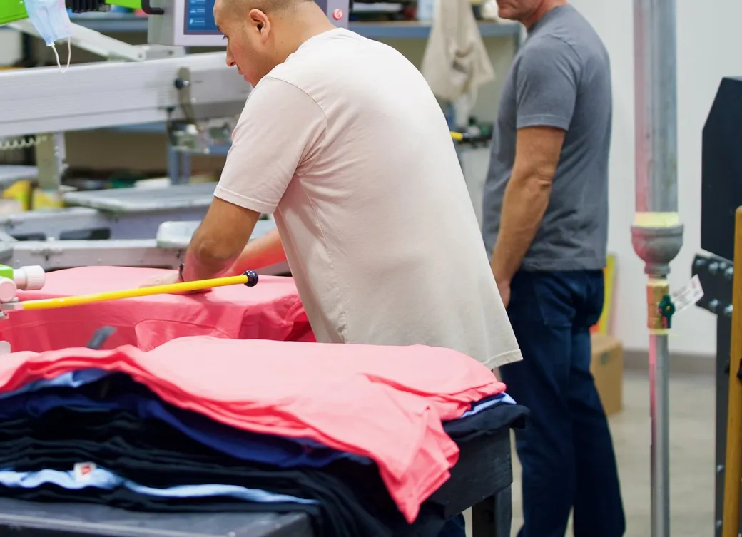Two printers working at the automatic press, with a stack of freshly printed coral tees on the table